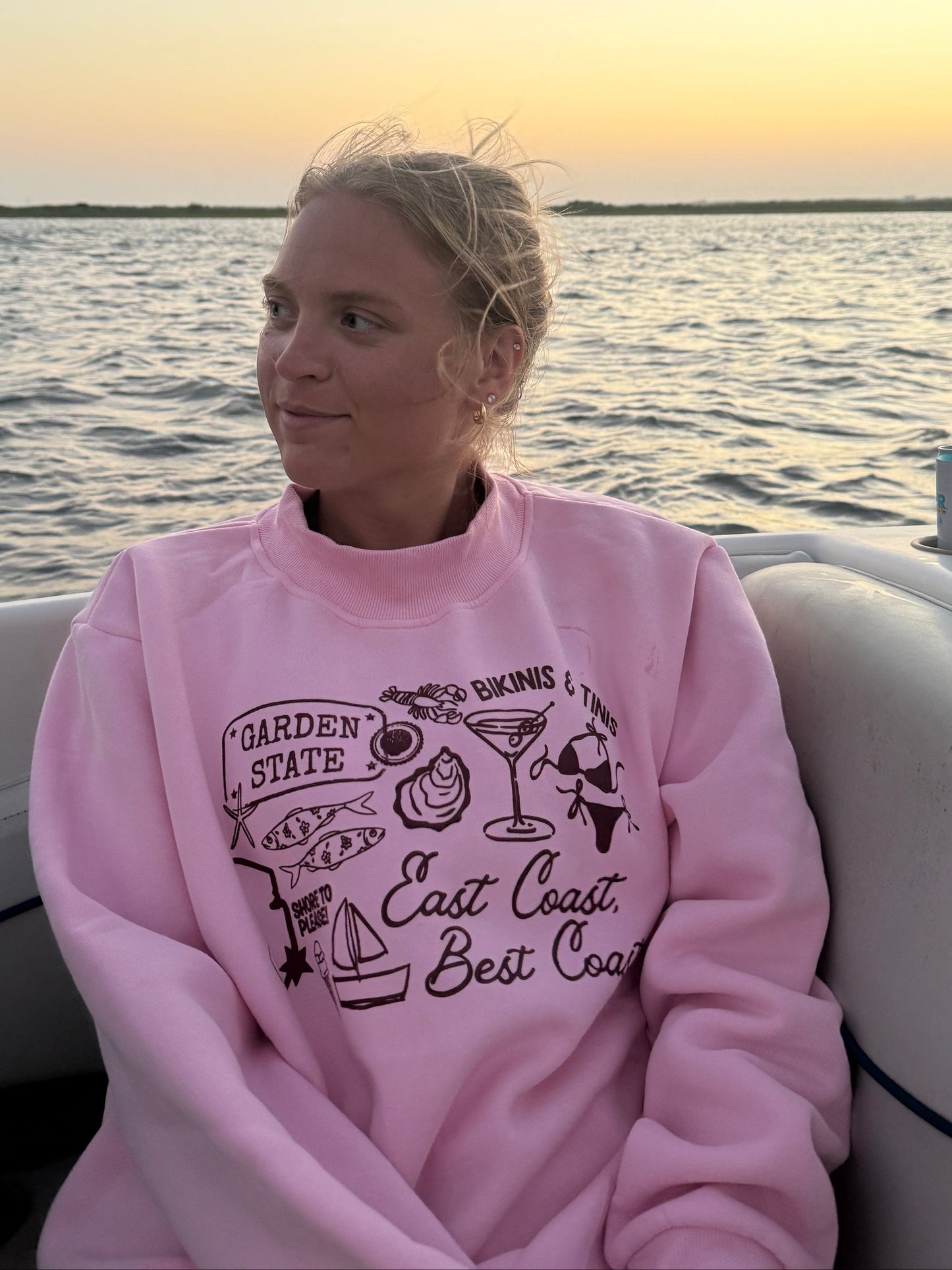 A woman wearing a pink long-sleeve jersey shore summer collage graphic mockneck sweatshirt sits on a boat with water and a sunset visible in the background